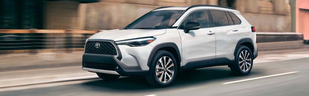 The image shows a white Toyota Corolla Cross SUV in motion on a city street, featuring a sleek design with a black grille and silver accents.