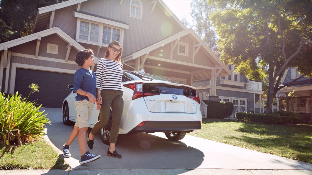 A woman and child walk in front of a white Toyota Prius, with a house in the background.
