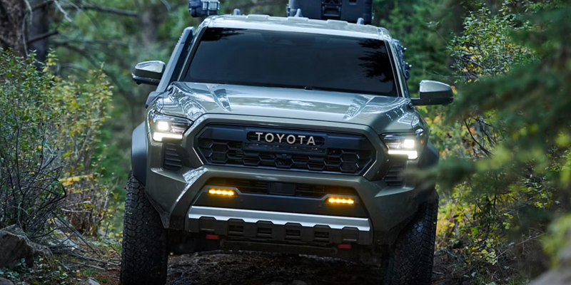 2026 Toyota Tacoma with bold grille, LED lighting, and off-road tires driving through a forest trail.