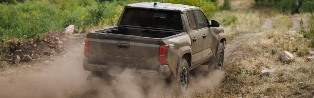 Dusty brown 2026 Toyota Tacoma pickup truck driving off-road on a rugged dirt trail, kicking up dust, with grass and rocks surrounding the path.