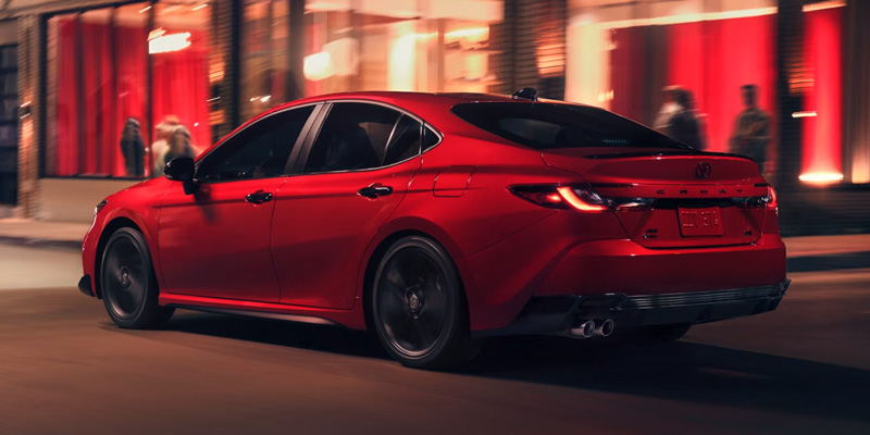 A red Toyota Camry Hybrid sedan parked on a city street at night with a blurred background of people and lights.