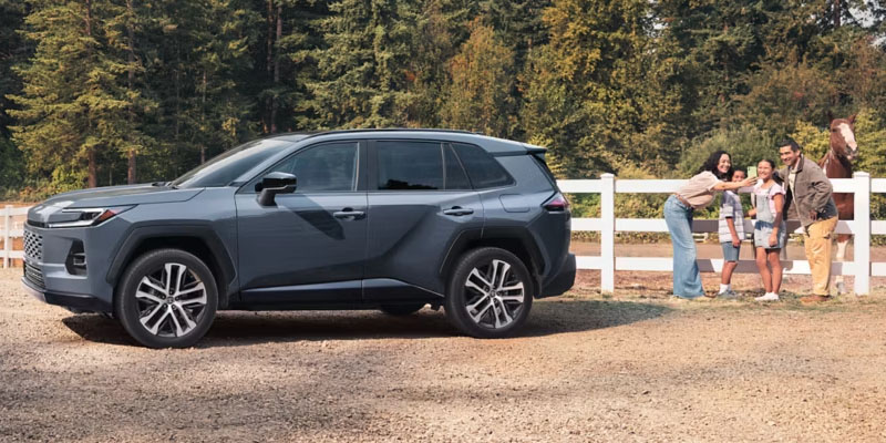 Gray Toyota RAV4 SUV parked on a gravel road beside a white fence, with a family standing near a horse in a wooded rural setting.