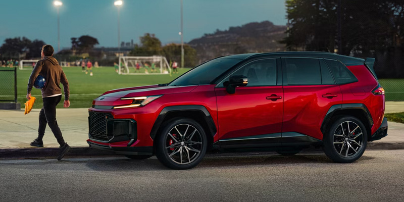 A red Toyota RAV4 Hybrid SUV parked on a street next to a soccer field, with a person walking away holding a helmet and a bag.