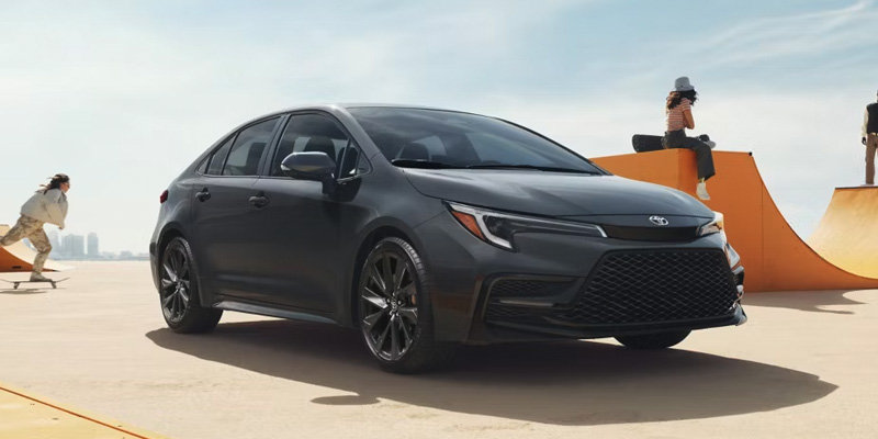 Black Toyota Corolla sedan parked in front of orange skate ramps, with skateboarders riding and relaxing in the background under a clear daytime sky.