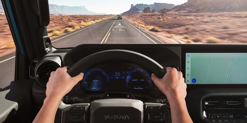 A person driving a Toyota vehicle on a deserted road with desert landscape and mountains in the background. The dashboard display shows the speed and navigation information.