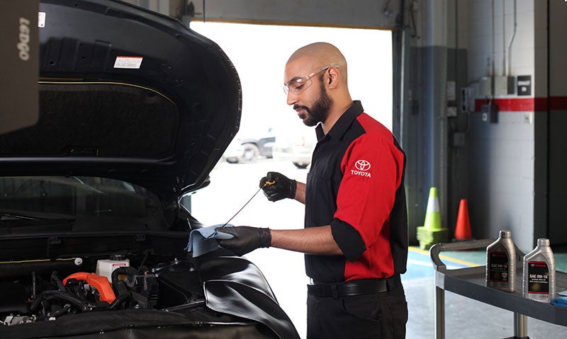 A mechanic wearing a Toyota uniform is checking the oil level in a car engine with a dipstick while standing next to an open hood.